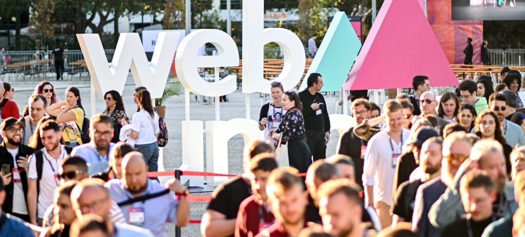 Pessoas no Web Summit 2023. A foto tem cores vibrantes, é um dia de sol. No fundo, vê-se o logo do evento, que consiste em dois triângulos, um menor, à esquerda, azul; outro maior, à direita, rosa. A imagem é usada para ilustrar uma matéria sobre Inteligência Artificial generativa, pois o tema foi debatido no Web Summit