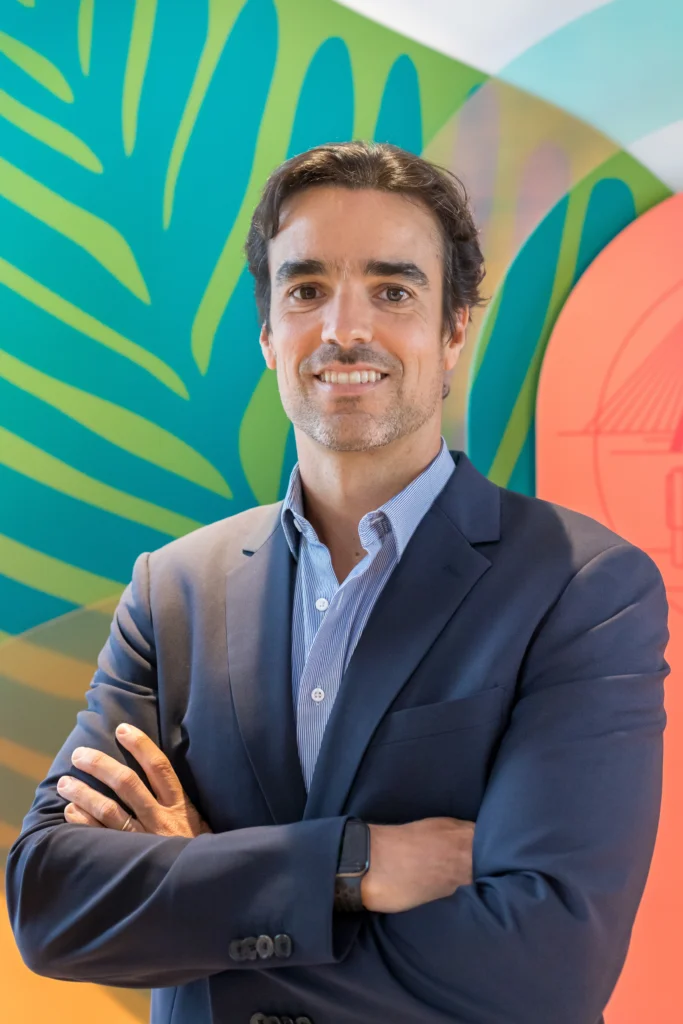 André Loureiro, diretor executivo do Pinterest  para a América Latina, posa de braços cruzados em frete à uma parede com uma pintura de folhas nas cores azul, verde e rosa. André usa um paletó azul escuro e uma camisa azul clara. 