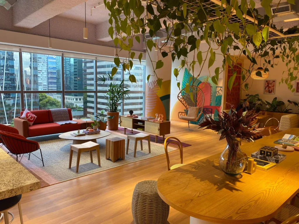Escritório do Pinterest em São Paulo. A iamgem mostra uma ampla sala com janelas grandes de vidro. É possível ver uma mesa de madeira com um vaso de plantas e diversos itens de decoração. Há um sofá vermelho, uma mesa de centro, bancos de madeira e uma poltrona vermelha. 