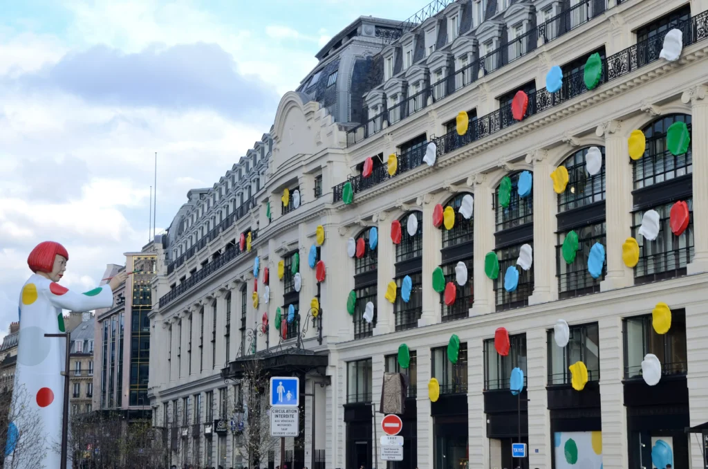 Uma estátua da artista Yayoi Kusama posicionada em frente à loja da marca Louis Vuitton, em Paris, França. A estátua mostra a artista com o cabelo curto vermelho e um vestido longo e de mangas compridas com bolas coloridas nas cores vermelho, azul, cinza, amarelo e verde. O prédio da loja da Louis Vuitton também está decorada com bolas coloridas. 