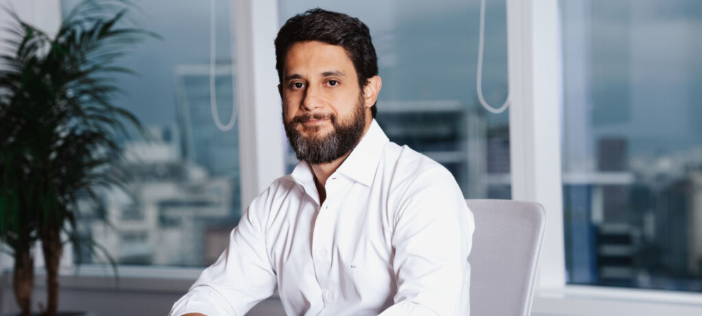 Marcelo Godoy veste uma camisa branca. Ele está sentado em uma cadeira. Ao fundo, está uma janela com vista para uma cidade, e uma planta à sua direita.