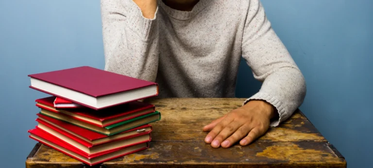 Uma pilha de livros com capas em diferentes tons de vermelho estão em cima de uma mesa de madeira. Atrás, está um homem de camiseta cinza com mangas longas. Ele apoia uma das mãos sobre a mesa. Atrás, uma parede azul.