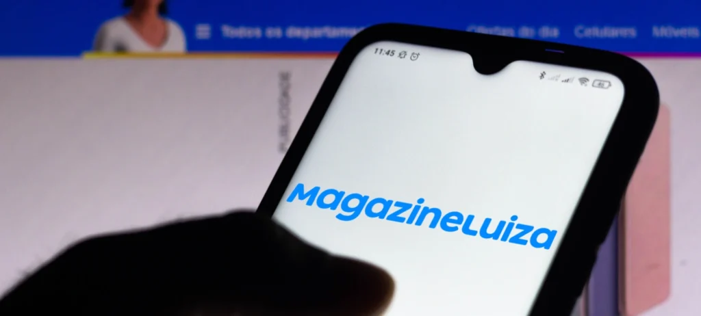 Uma pessoa utiliza o celular para acessar o aplicativo da Magazine Luiza no celular. A tela do celular está em branco com as letras em azul escrito "Magazine Luiza". Ao fundo, há a tela de um computador navegando no site da Magalu.