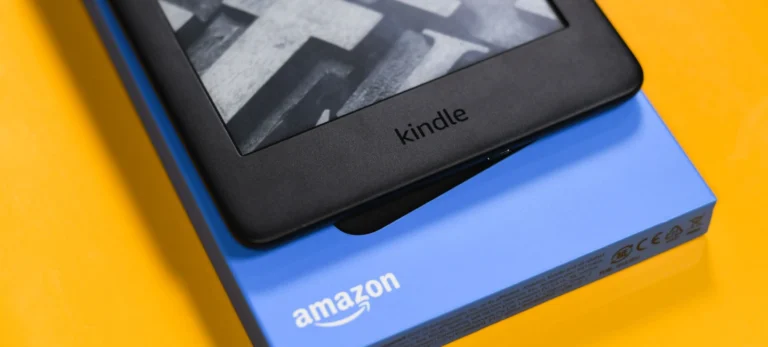 Um aparelho Kindle é colocado sobre uma caixa azul com o logotipo da Amazon, com um fundo amarelo.