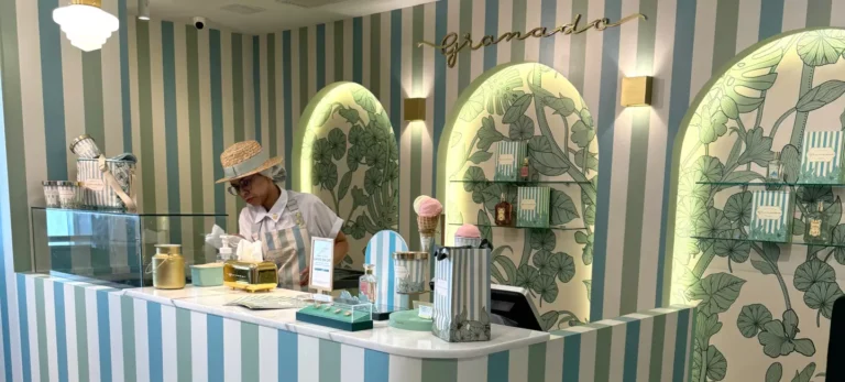 A foto mostra o interior da loja da Sorveteria Granado Verão. O espaço tem paredes listradas verde claro e branco. Uma bancada expõe as variedades de sorvetes da loja. Uma pessoa atrás do balcão serve um sorvete.