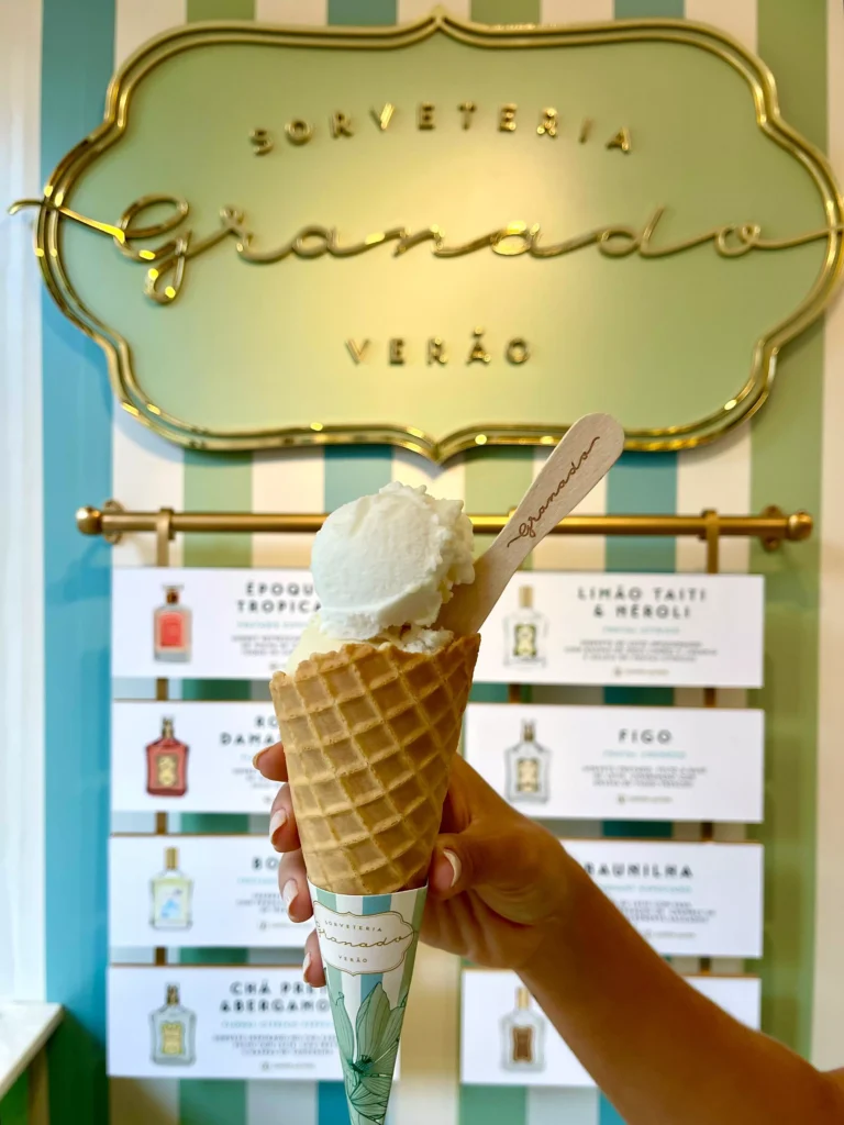 Um sorvete no cone é segurado por uma mão. Atrás, há um cardápio com as opções de sorvete da Sorveteria Granado Verão, e uma placa com o nome da loja. A parede é listrada nas cores verde, azul e branco. 