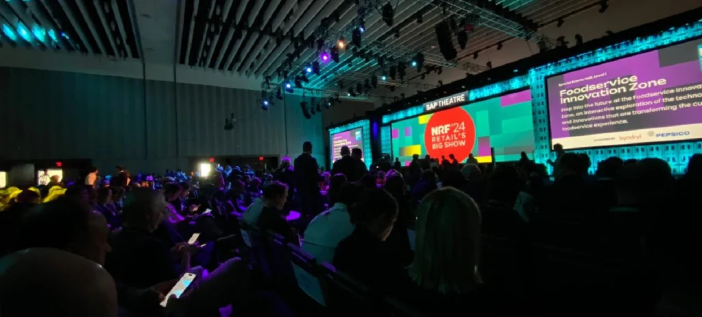Plateia da NRF acompanha painel durante evento. As luzes estão apagadas, e apenas o telão está iluminado. Algumas pessoas utilizar os celulares.