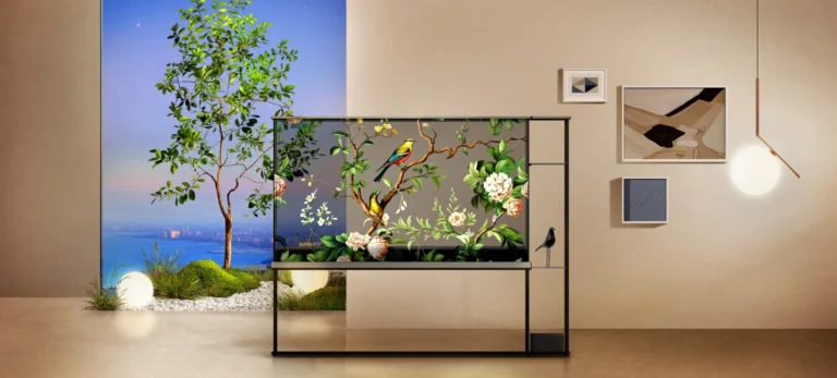 O modelo de televisão OLED Signature T, da LG Eletronics, está em uma estante de metal no meio de uma sala com piso de madeira e parede bege. Atrás da televisão, está um quadro com a foto de uma árvore, outros pequenos quadros e uma luminária.