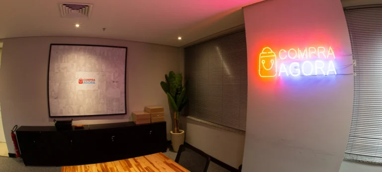 Foto mostra uma sala de escritório do Compra Agora. À direita, um letreiro em neon vermelho e azul mostra o logo da empresa. À esquerda, um móvel preto baixo e um quadro com a logo do Compra Agora.