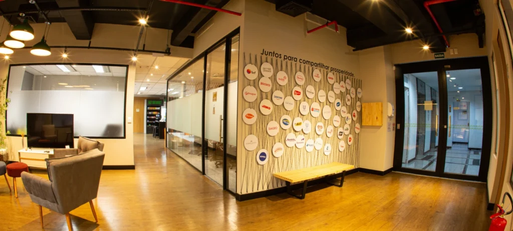 Foto mostra a entrada do escritório do Compra Agora. Em uma das paredes, um painel com as logos de todas as indústrias que fazem parte do ecossistema. À esquerda, poltronas, mesas e uma sala de reunião. 