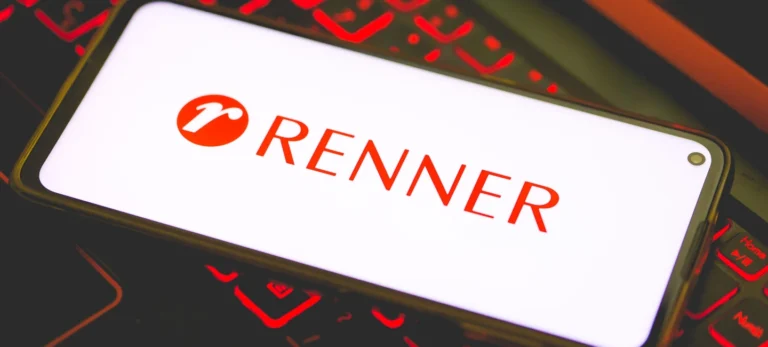 Um smartphone mostra a imagem da logo das Lojas Renner em vermelho sobre um fundo branco. Abaixo do celular, um teclado de notebook com luzes vermelhas ao redor das teclas.