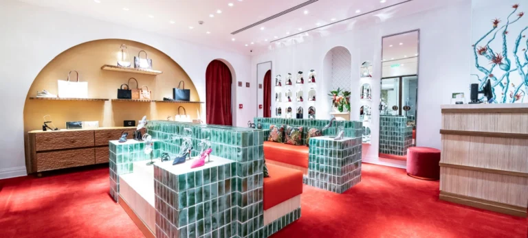 A foto mostra a nova loja de Christian Louboutin em São Paulo. Sofás vermelhos possuem armação coberta por azulejos verdes. A parede branca possui prateleiras com sapatos e bolsas da marca. O carpete é vemelho.