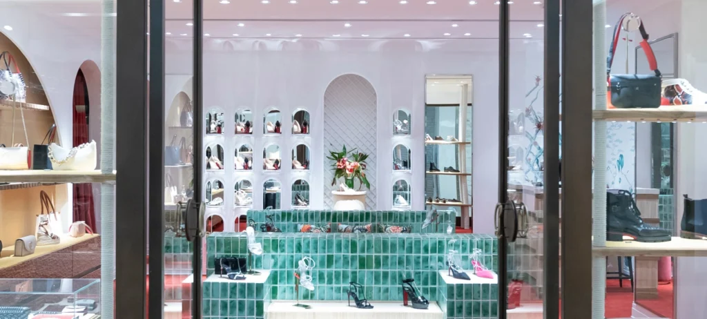 Foto mostra a loja de Christian Louboutin em São Paulo. Na vitrine, prateleiras estão repletas de bolsas e sapatos de diferentes formatos e cores. No interior da loja, uma bancada com azulejos verdes apresenta outros produtos. Na parede branca ao fundo, um vaso de flores e mais produtos da marca. 