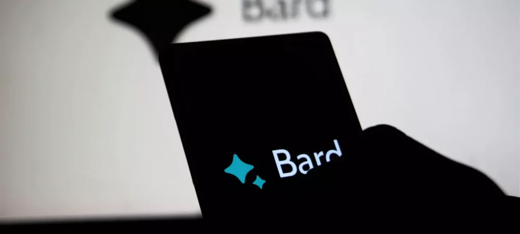 Uma pessoa segura um celular que exibe a logo tipo do Bard, IA generativa do Google, sobre um fundo preto. Atrás, há uma tela de computador que também exibe o logo sobre um fundo branco.