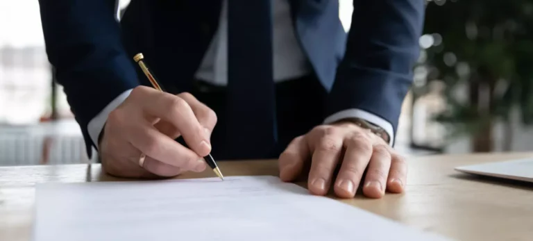 Um homem vestido de terno assina um documento, apoiado sobre uma mesa.