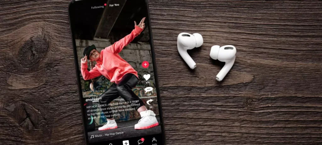 Um celular está aberto no TikTok e reproduz um vídeo de um homem de casaco vermelho dançando. Ao lado do celular, há um par de fones de ouvido sem fio.