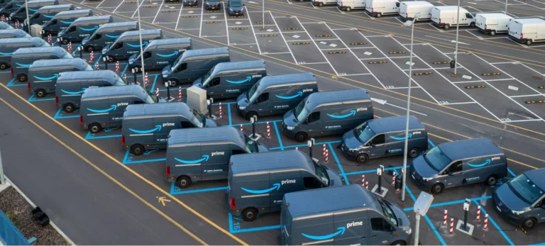Foto mostra estacionamento com várias vans elétricas de entrega da Amazon Prime de cor azul escuro.