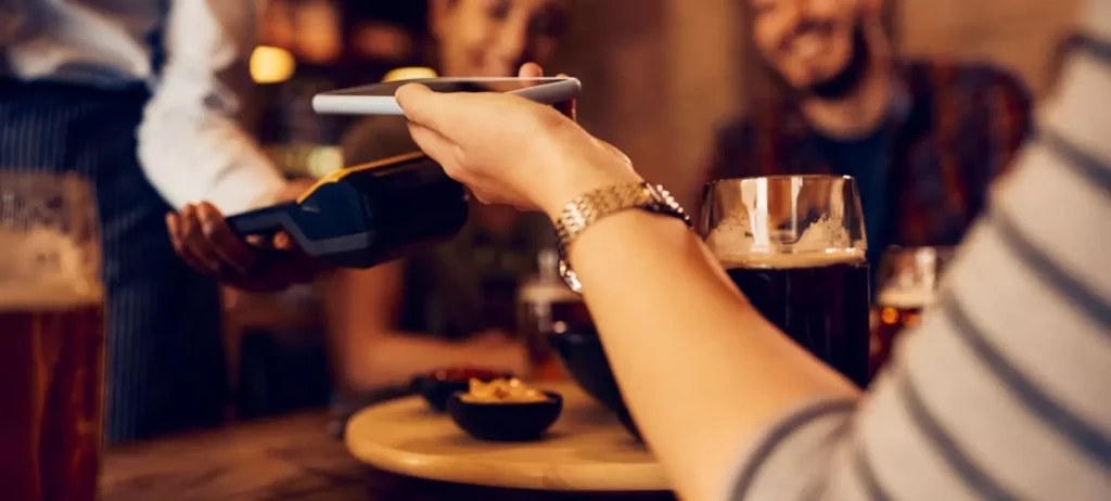 Uma mulher em um restaurante paga pela conta da refeição utilizando o celular e aproximando-o na maquininha.