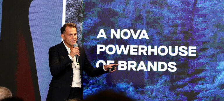 Alexandre Birman, CEO da Arezzo, em apresentação a investidores. Atrás dele, um telão exibe a frase "a nova powerhouse of brands" em branco sobre um fundo azul.