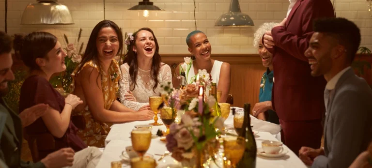 Foto mostra um casamento intimista. A noiva é cercada por três mulheres. Elas estão sentadas diante de uma mesa, repleta com copos e flores de diferentes cores. Um homem está de pé em frente a elas.