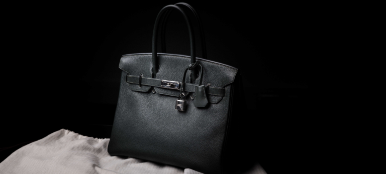 A foto mostra uma bolsa Birkin, da Hermès, preta sobre um pano cinza claro. Atrás, um fundo preto.