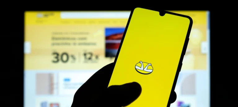 Uma mão segura um smartphone que exibe a logotipo do Mercado Livre: duas mãos que se apertam sobre um fundo amarelo.