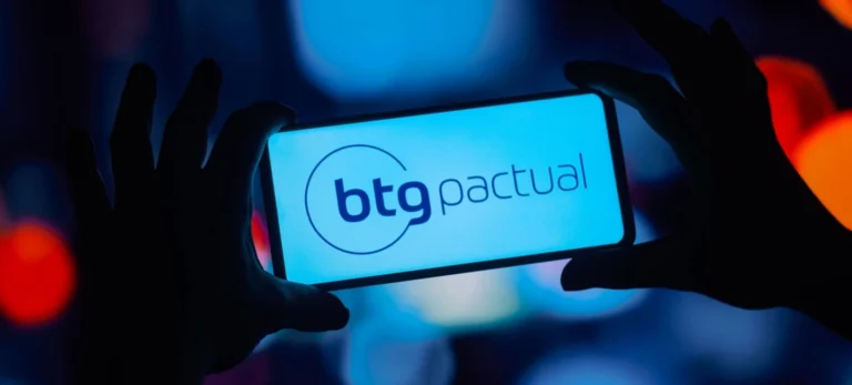 Imagem mostra um par de mãos de segurando um smartphone na sombra. O brilho da tela mostra a logotipo do banco BTG Pactual.