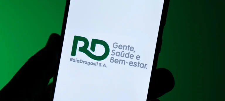 Foto mostra uma tela de celular com a logotipo do Grupo Raia Drogasil. Atrás do celular, há um fundo em um verde escuro.