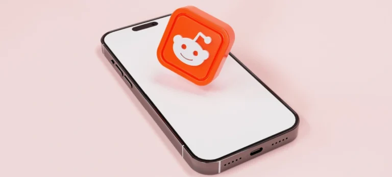 Um smartphone com a tela em branco exibe o logo do Reddit. O celular está apoiado sobre um fundo rosa claro.
