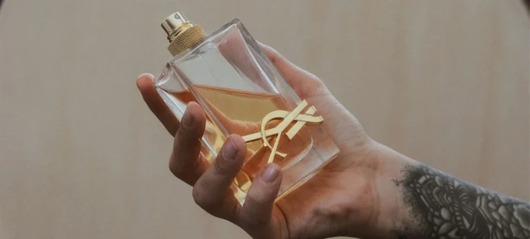 Uma mão com o pulso tatuado segura um frasco de perfume da Yves Saint Laurent.