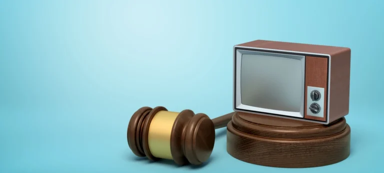Uma televisão vintage está apoiada sobre um apoio de madeira. Do lado, o martelo da justiça. O cenário é azul claro.
