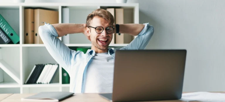 Um homem de cabelo loiro e curto e de óculos reclina em uma cadeira com as mãos atrás da cabeça. Sua expressão é de felicidade e alegria. À sua frente, está um computador e um caderno.