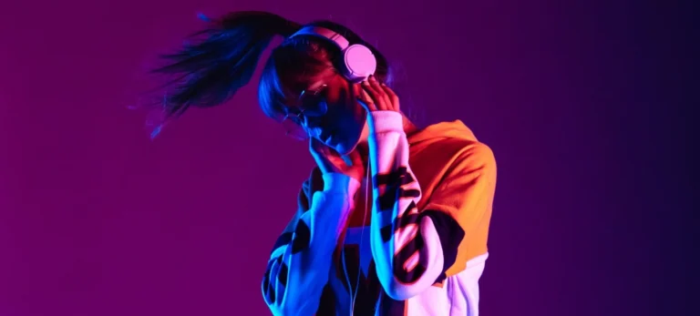 Uma jovem mulher ouve música por meio de um headset branco. Ela usa um moletom colorido e está com o cabelo preso em um rabo de cavalo.