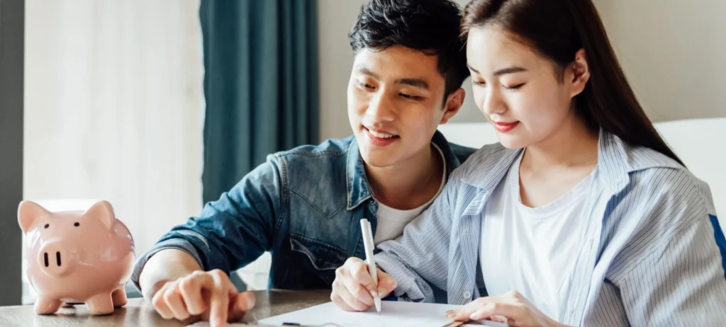 Um casal de jovens analisa uma pilha de papeis sobre uma mesa. Os dois sorriem. Ao lado, há um cofrinho no formato de um porco rosa.