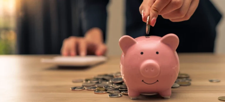 Uma pessoa deposita uma moeda em um cofrinho no formato de um porco cor de rosa claro.