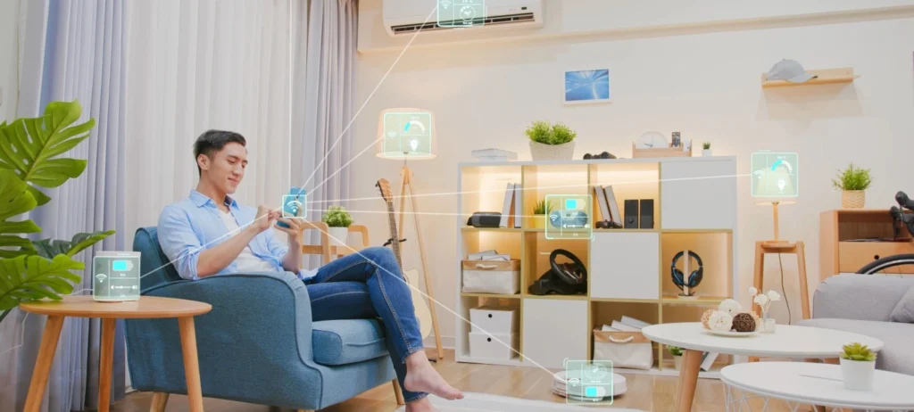 Um homem está sentado em uma cadeira na sala de estar. Ele utiliza um smartphone, conectado aos diferentes eletrônicos da casa.