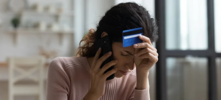 Uma mulher segura um cartão de crédito numa mão e o telefone na outra, com a aparência preocupada.