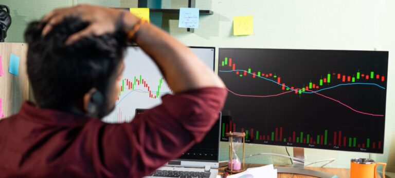 Um homem parece aflito, com as mãos na cabeça, enquanto observa um gráfico com uma seta vermelha em queda, na tela de um computador.