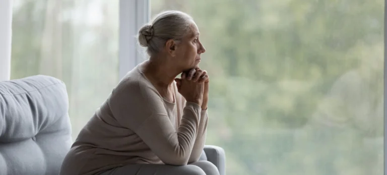 Uma mulher branca e de cabelos grisalhos preso em um coque olha pensativa através de uma janela de sua casa. Ela está sentada em um sofá cinza claro e veste roupas da mesma cor.