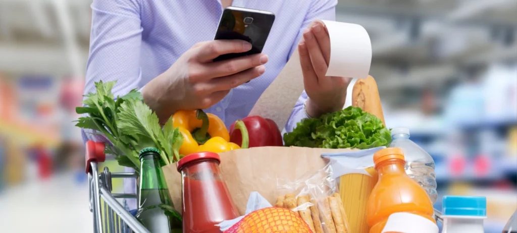 Uma mulher segura o celular e a nota fiscal de suas compras de mercado.
