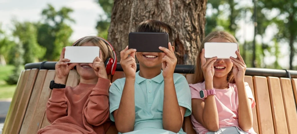 Três crianças estão sentadas em um banco numa praça, e cada uma segura um smartphone na altura dos olhos, tampando seus rostos.