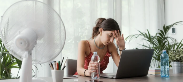 Uma jovem mulher parece assolada pelo calor, e tem um ventilador ligado sobre sua mesa.