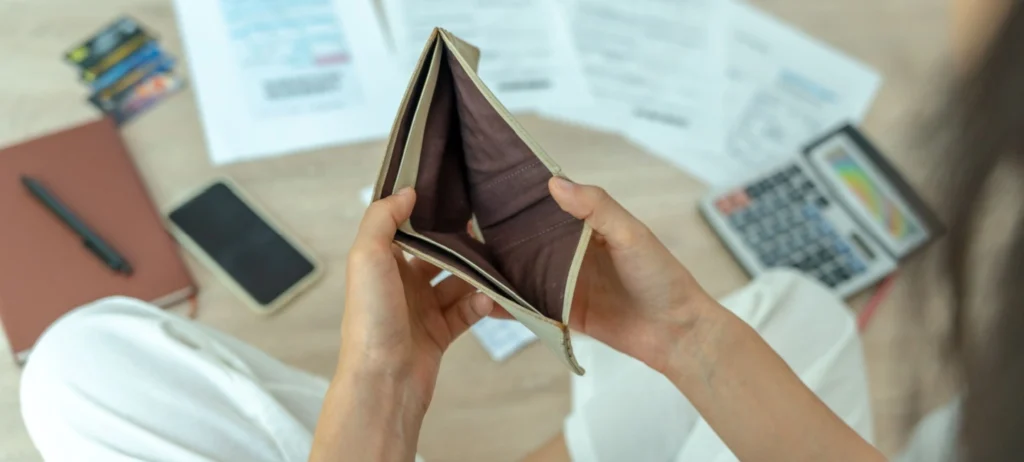 Uma pessoa segura uma carteira aberta vazia, e no chão, há várias contas em papel, além de um bloco de anotações.