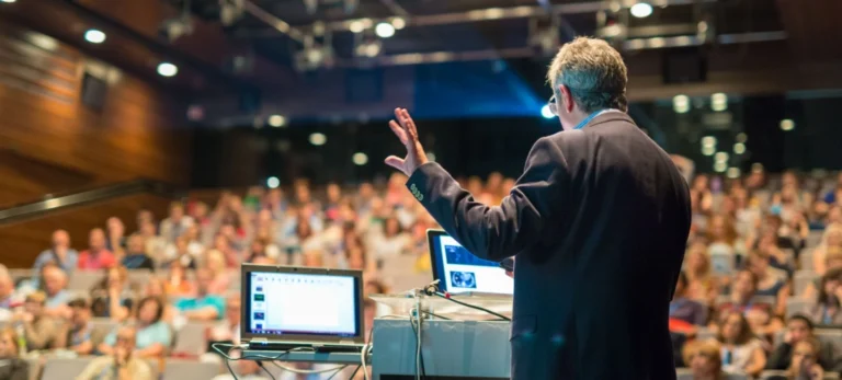 Um homem está de costas, palestrando para uma plateia em um auditório.