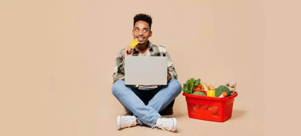 Um homem está sentado no chão, segurando um notebook no colo. Ao seu lado, uma cesta vermelha está cheia de produtos de alimentos.