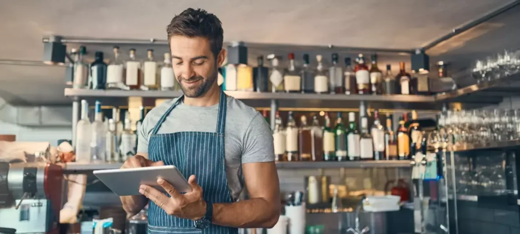 Um homem usa um avental e mexe em um tablet. Ele está em uma cafeteria ou restaurante.