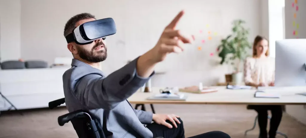 Um homem está sentado em uma cadeira de rodas. Ele usa um óculos de realidade virtual e aponta o dedo para cima.