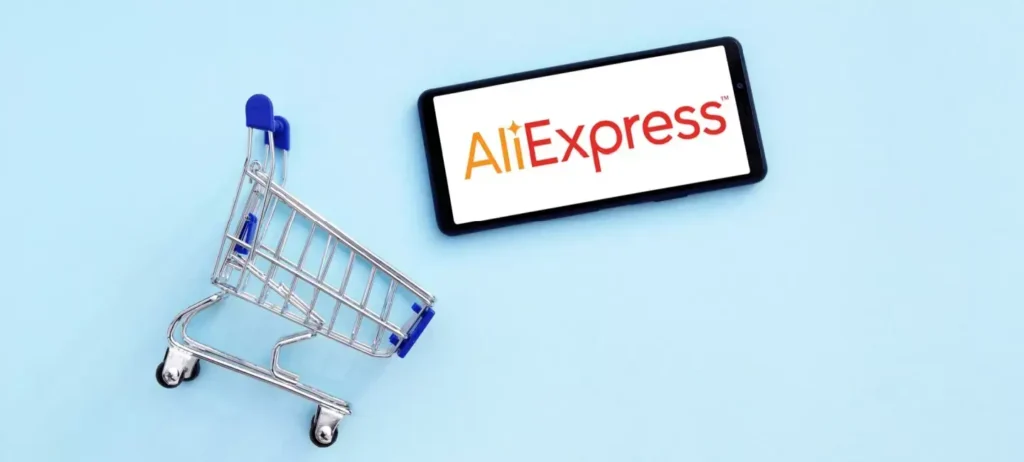 Um carrinho de compras está deitado no chão. Ao lado, um smartphone mostra a logo do AliExpress.