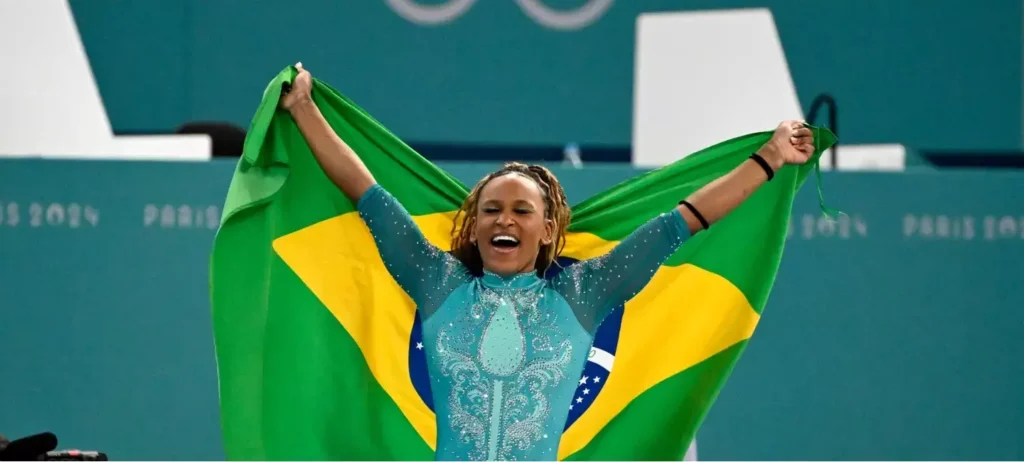 A ginasta Rebeca Andrade ergue a bandeira do Brasil em celebração à conquista da medalha de bronze nas Olimpíadas de Paris.