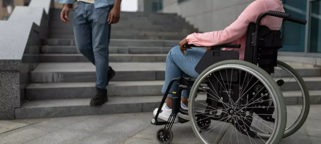 A imagem mostra uma jovem mulher usando uma cadeira de rodas. Ela está diante de uma escada, enquanto por ela desce um jovem rapaz.
